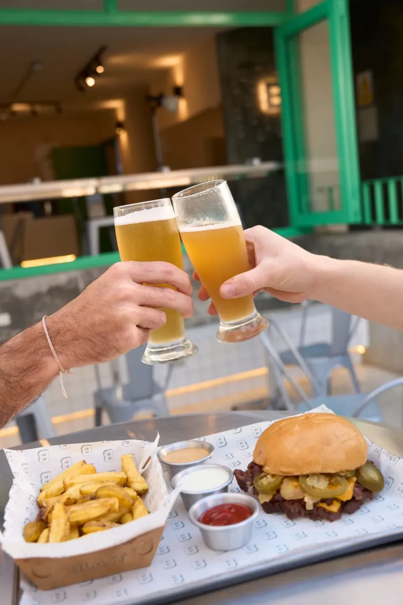Dos personas brindando con cerveza sobre una mesa con burgers y papas de Blast