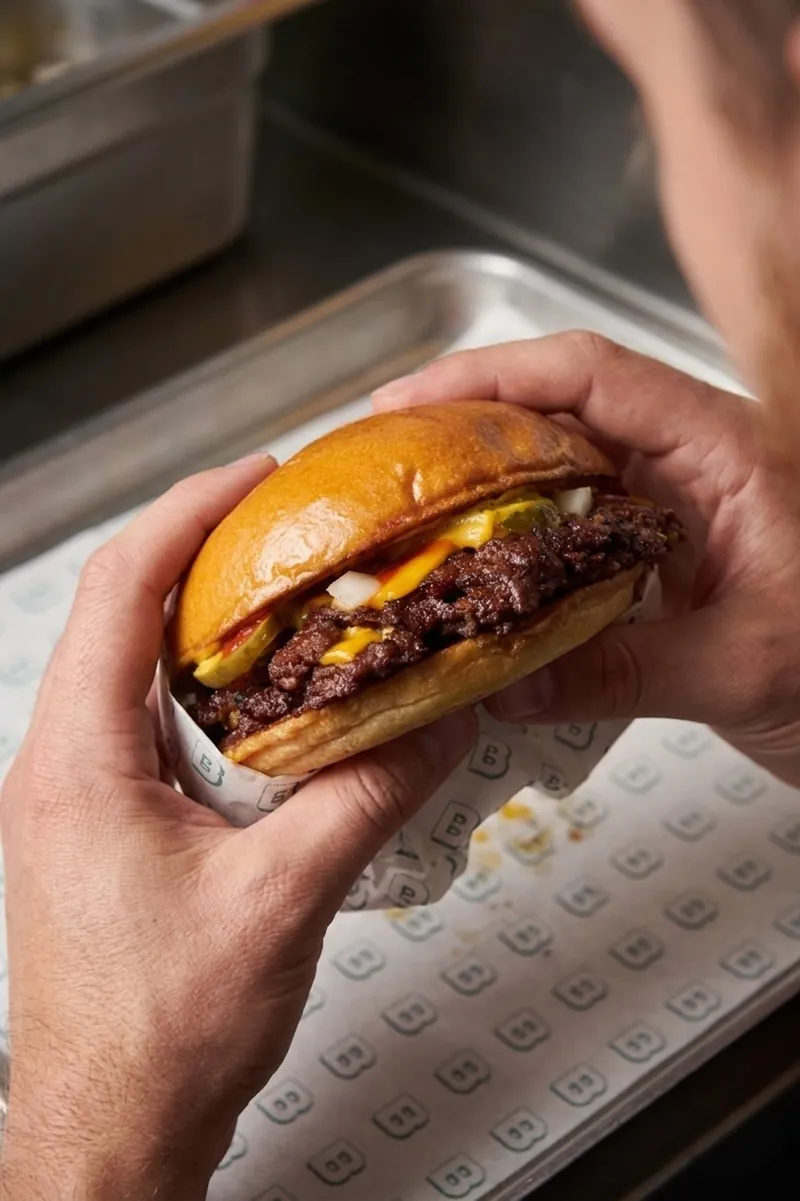 Manos sosteniendo una smash burger Blast con queso fundido y bordes caramelizados