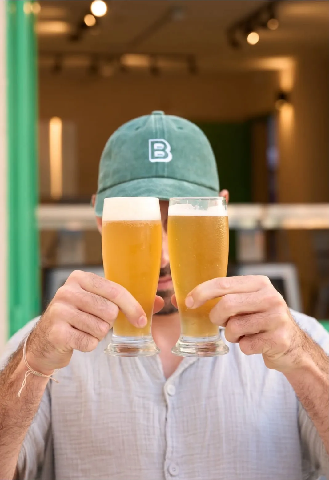 Persona con gorra verde de Blast sosteniendo dos vasos de cerveza artesanal rubia frente a su cara